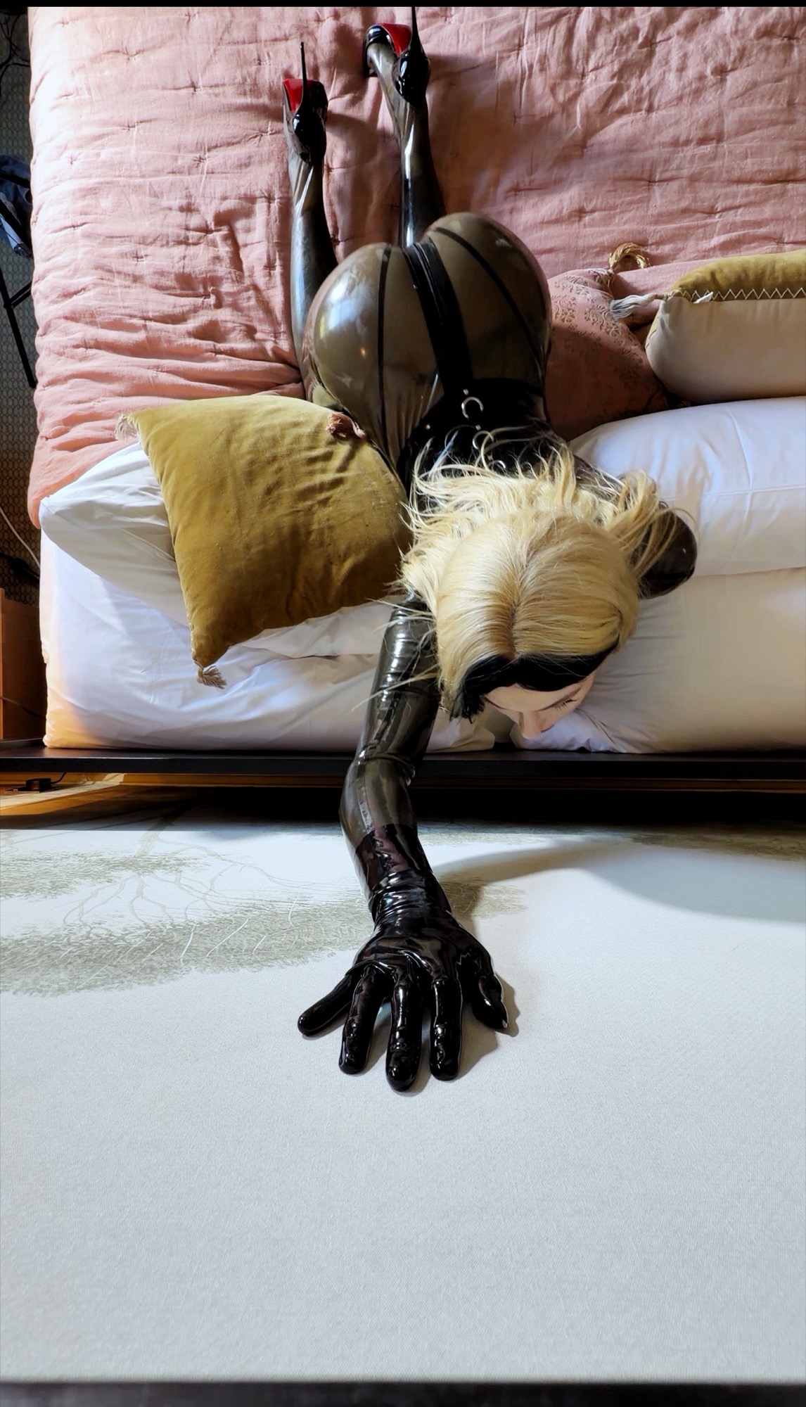 Aria face down across a bed in a black latex catsuit and heels, one gloved hand reaching to the floor.