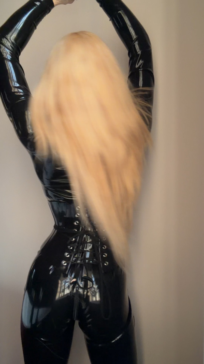 Aria from behind in a laced black latex catsuit, long blonde hair falling down her back.